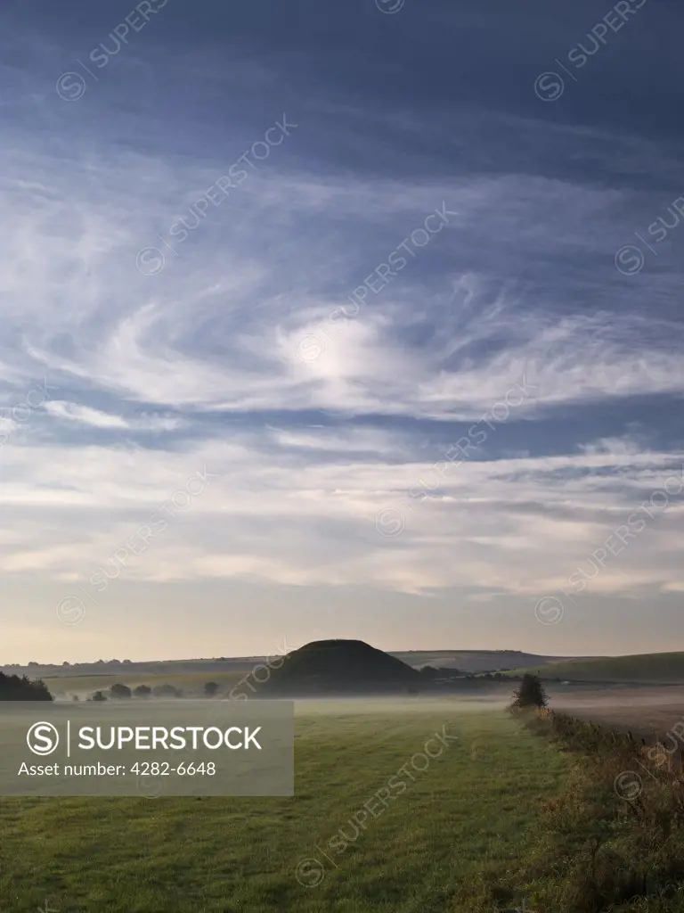 England, Wiltshire, Near Avebury. View across misty field to Silbury Hill, the tallest prehistoric human-made mound in Europe, beneath swirling clouds.