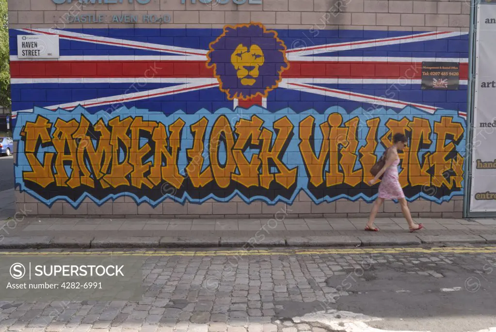 England, London, Camden. A woman walking past a painting on a wall in Haven Street, Camden, featuring the head of a lion above the words Camden Lock Village, over a union flag.