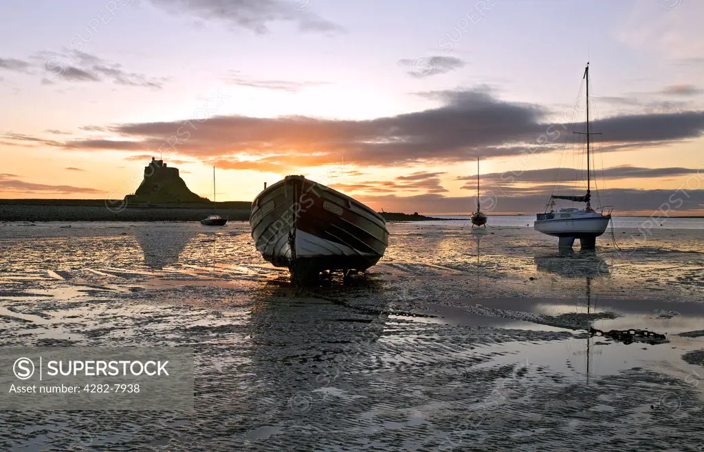 England, Northumberland, Lindisfarne. Sunrise over boats moored at low tide with a view to the castle at Lindisfarne.