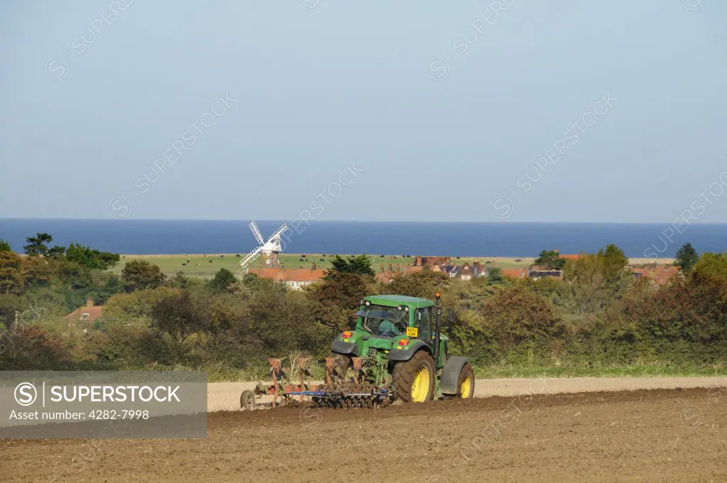 England, Norfolk, Weybourne. Tractor with combined plough and disc harrow on coastal farmland with Cley windmill in the distance.