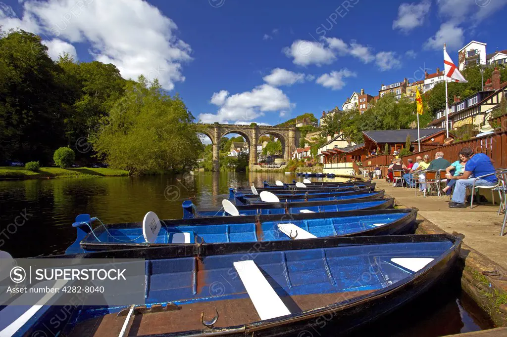 England, North Yorkshire, Knaresborough. Rowing boats for hire and people enjoying a drink at a cafe on the riverside of the River Nidd at Knaresborough.