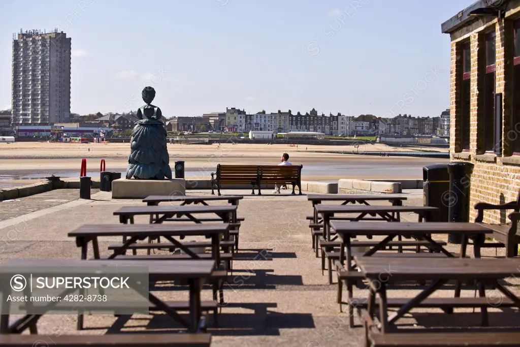 England, Kent, Margate. Trestle tables outside the Lighthouse bar. A nine feet high bronze sculpture of Mrs Booth, the Shell Lady of Margate by Ann Carrington, stands at the end of the Harbour Arm.