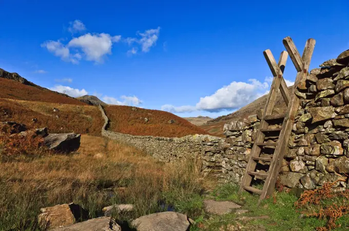 England, Cumbria, Derwent. A stone wall leads from a wooden stile to the hills beyond.