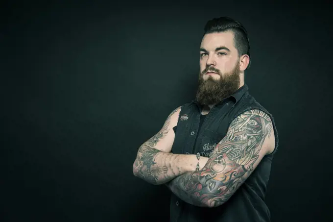 Studio portrait of a bearded man with tattooed arms.