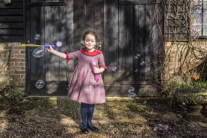 A six year old girl blowing bubbles by a garage door.