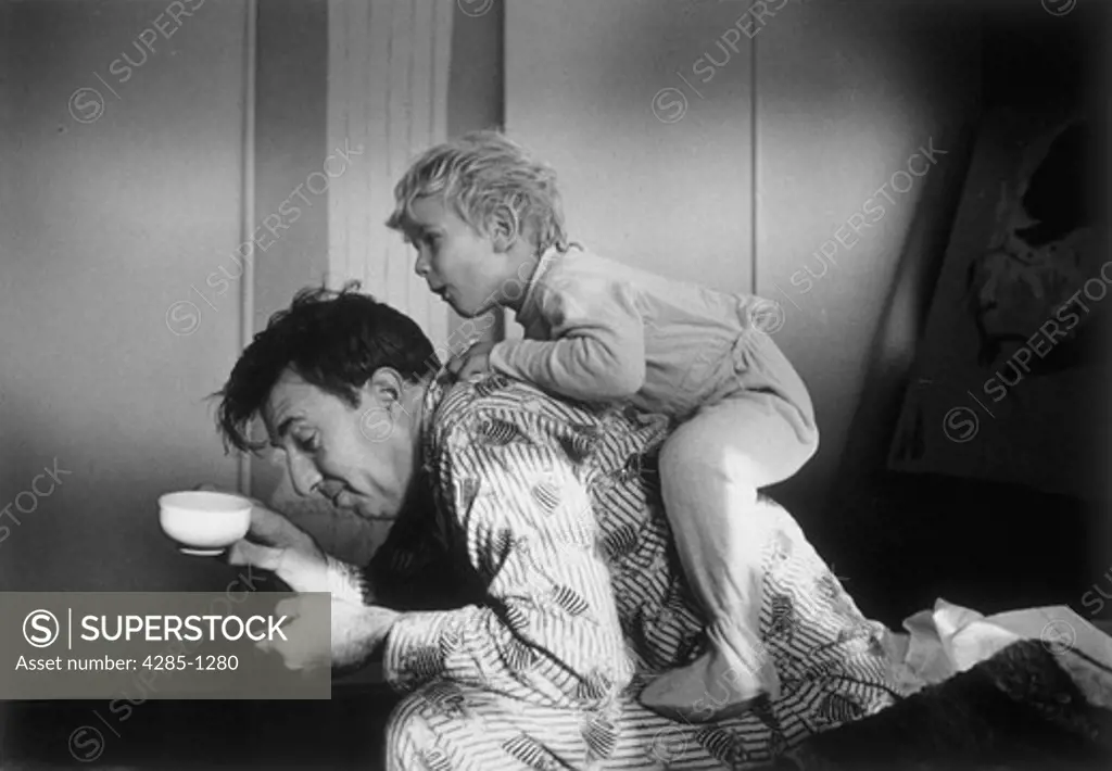 Eager young son jumps on his fathers back while his father tries to drink a cup of coffee in the morning.