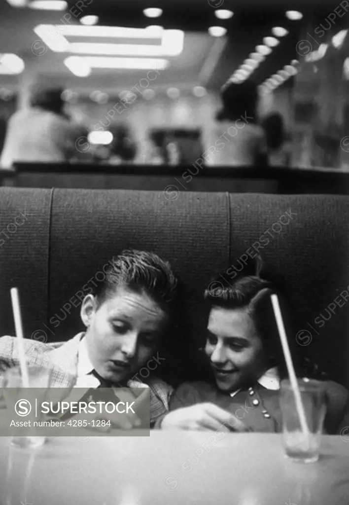 Teenage boy and girl sitting in a booth at a 1950s drugstore sharing soda on a date.