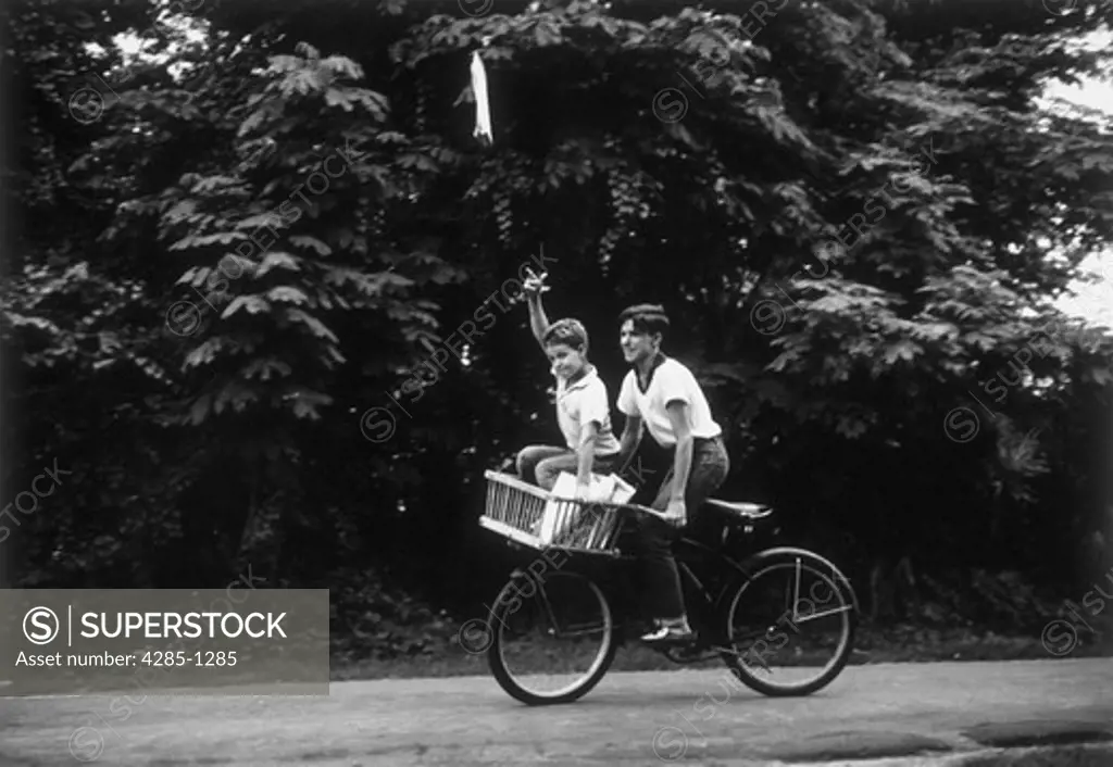 Two young brothers working together to deliver newspapers.  One boy pedals the bicycle while the other rides on the handle bars and throws the papers.