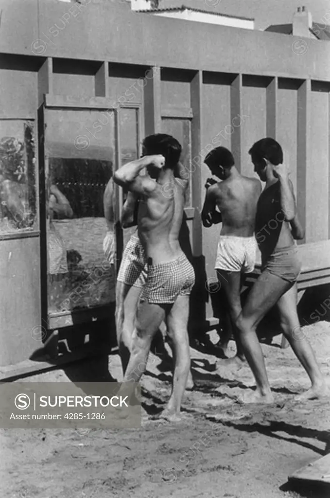 Three teenage boys flexing their muscles and posing in front of mirrors outdoors at the beach.   Image was taken in the 1950s.