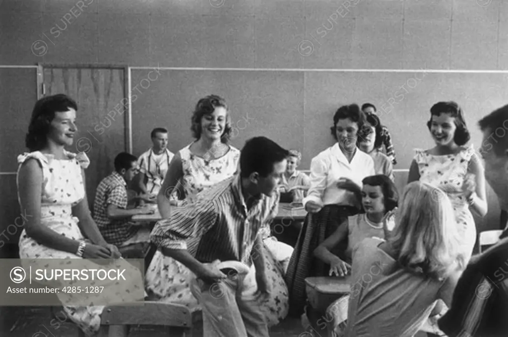 Classroom full of teenage boys and girls tease and flirt with eachother while the teacher is out of the room.   Image was taken in the 1950s.