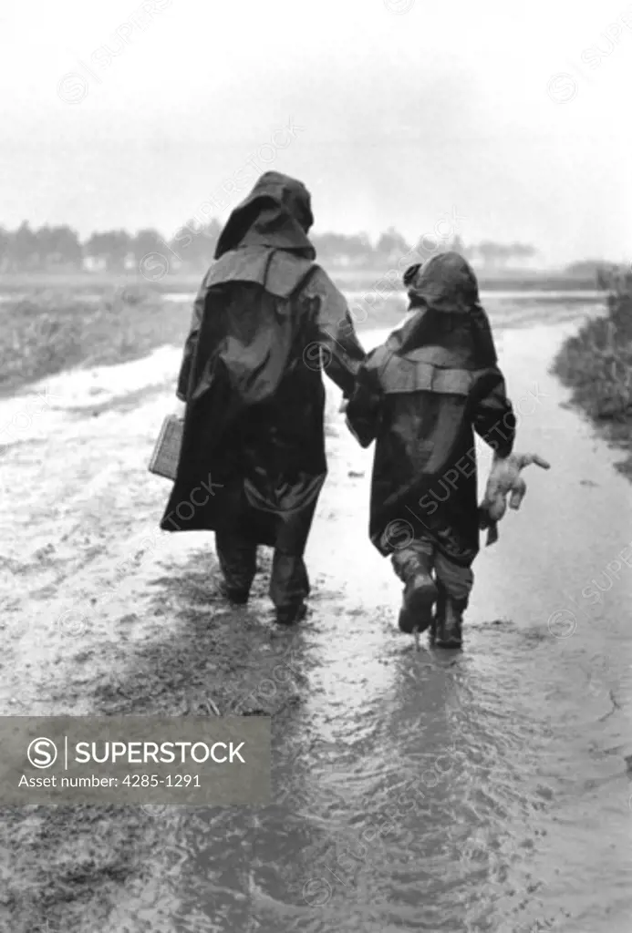 Two children in rain hats and raincoats walking in the rain.