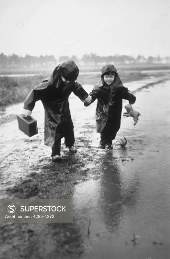Two young brothers wearing dark rain slickers jump in puddles while walking to school together.