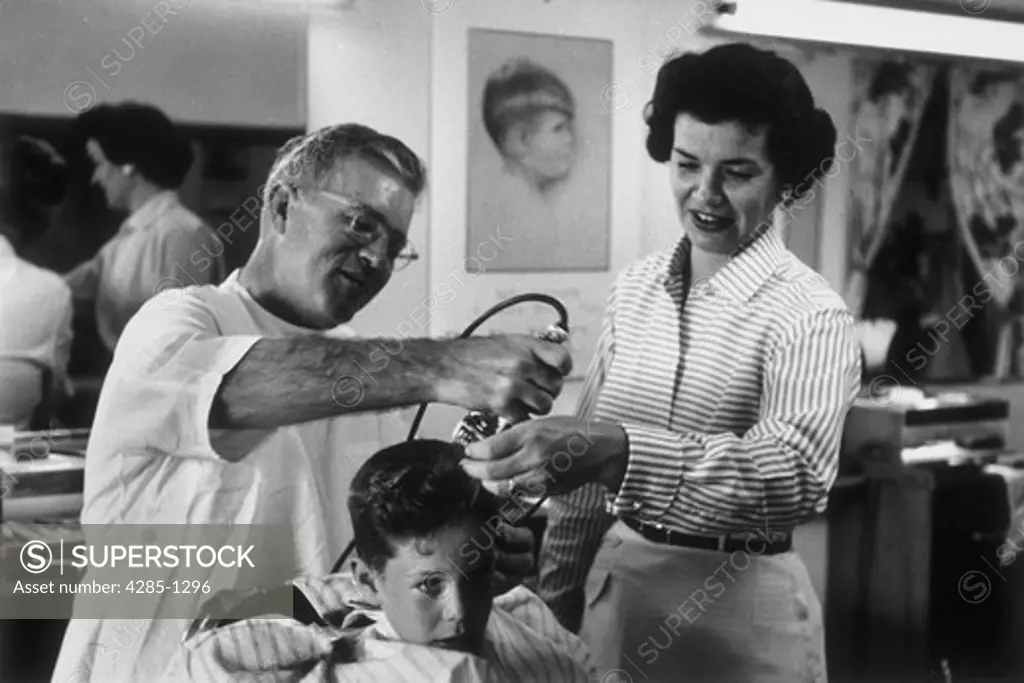 Young boy getting his first haircut from a barber while his mother looks on.   Image was taken in the 1950s.