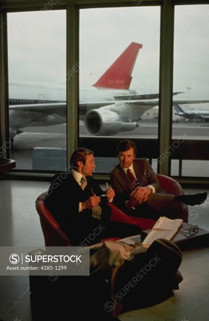 Two businessmen talking in lobby at airport with aiplane through window in the background.