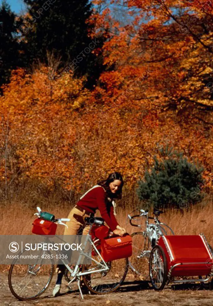 Woman preparing for bicycle camping trip by packing bicycle pouches outdoors with fall foliage.