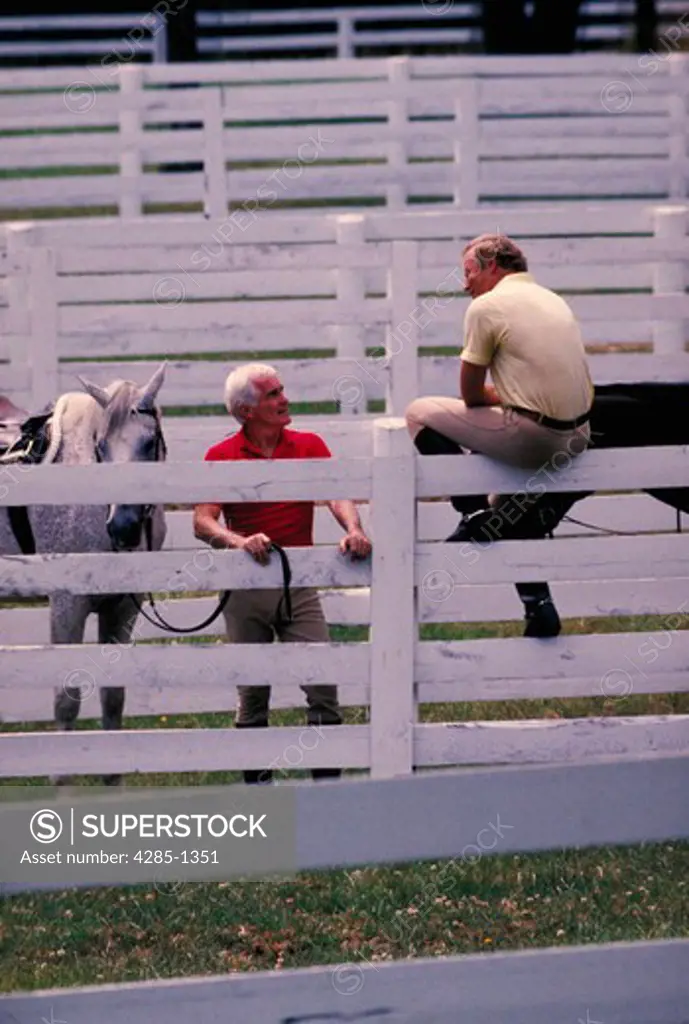 Two men with horses talking over white fence.