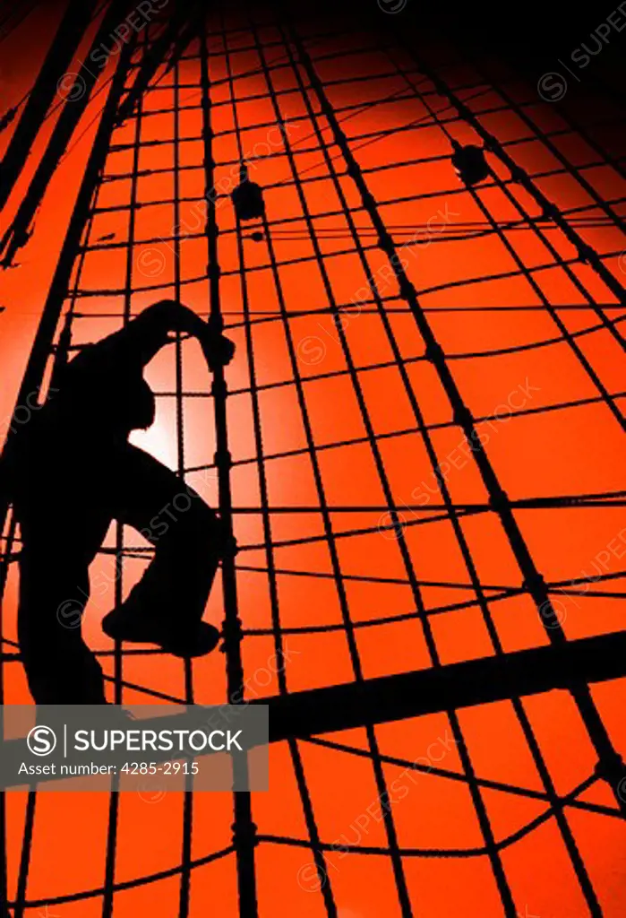Silhouette of a man climbing the riggings on the mast of a sail boat.