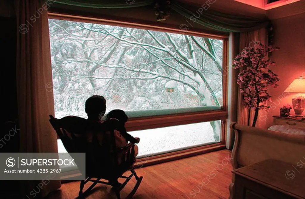 A man sitting with his dog in a rocking chair in front of a large picture window looking out at snow covered trees.