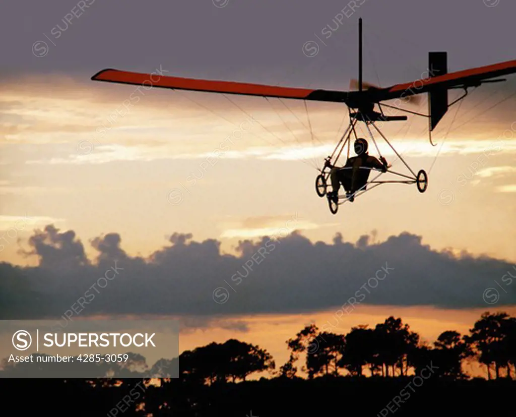 Ultralite flyer in silhouette with trees in sunset background.