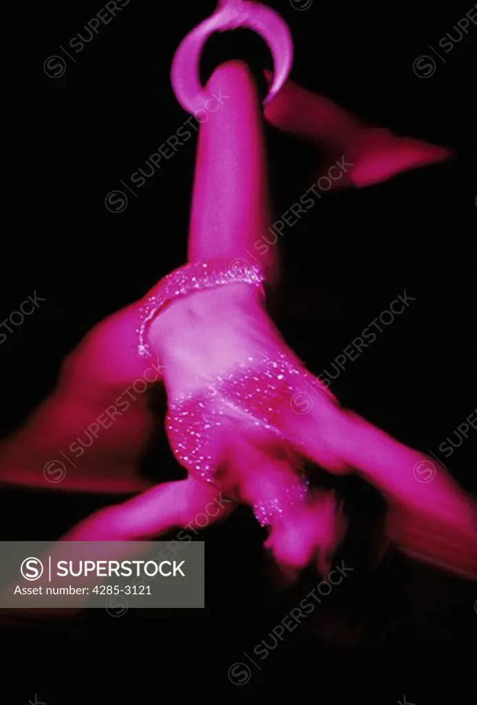 Woman's blurred figure bathed in magenta light, black background., (in circus show) suspended upside down by one leg in ring attached to high-wire.