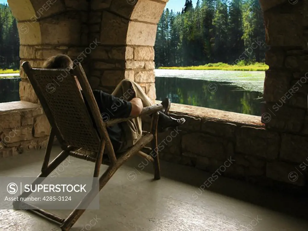 Man relaxing on lounge chair, seen from behind, in front of opening in rock porch, with scenic and placid green lake and pine trees in background.