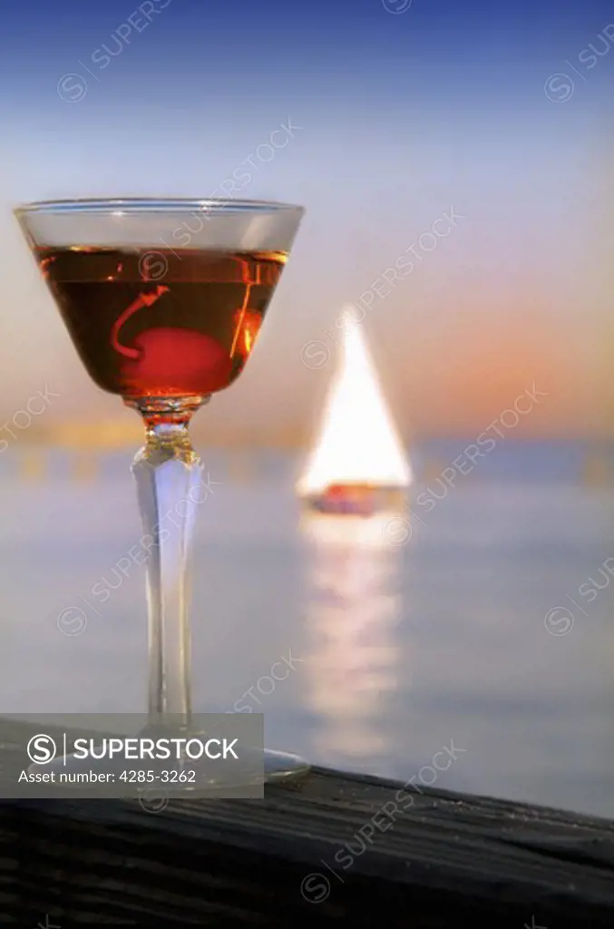 Vermouth drink and glass close-up, cherry inside, on wood beam with fiery sunset and sailboat in ocean in background.