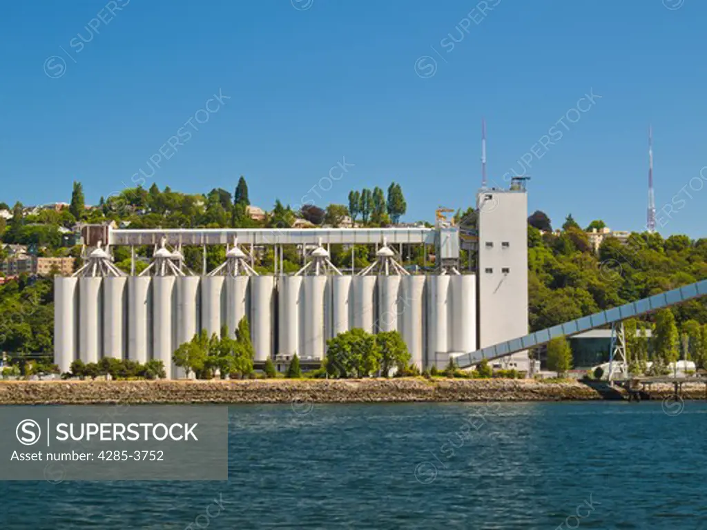 grain silos in a row by water's edge with houses on a hill behind it