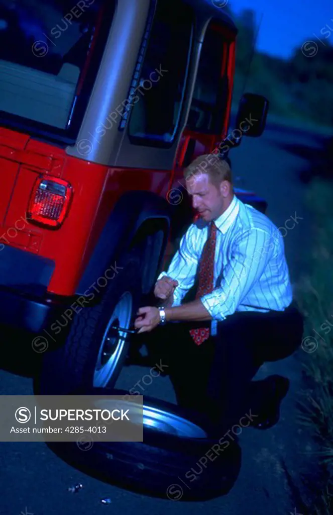 Businessman with his shirt sleeves rolled up changing a tire on his car on the side of the highway.