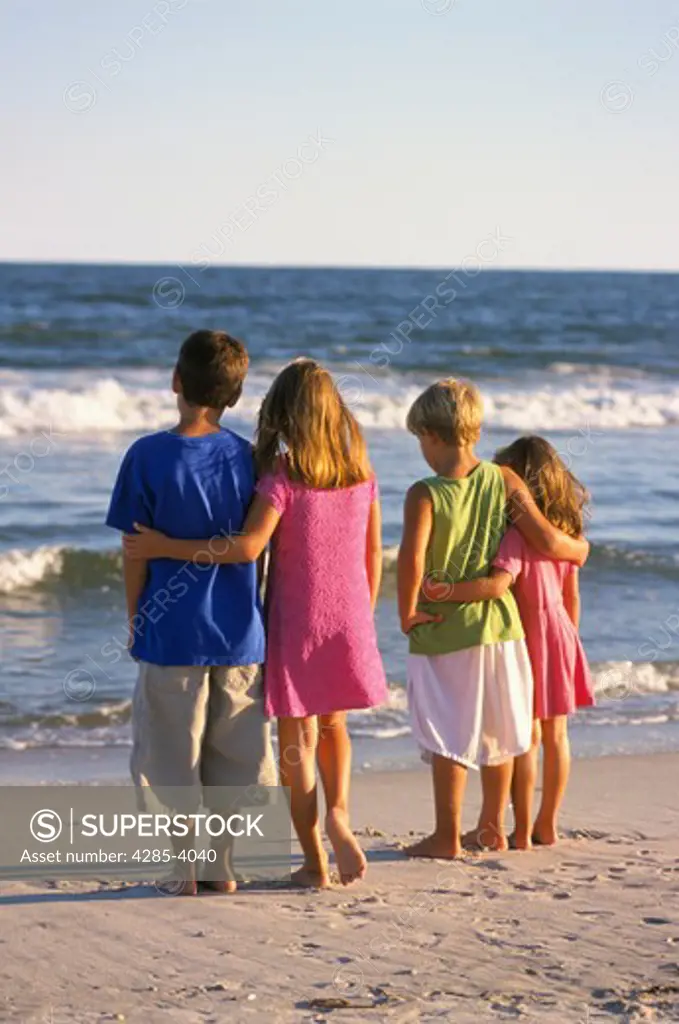 Four children on a beach standing quietly together watching the gentle waves break on shore.