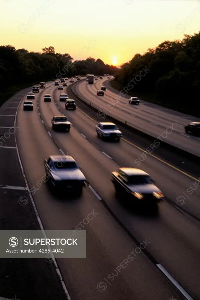 Cars traveling on a freeway in the early evening as the yellow sun sets behind them.
