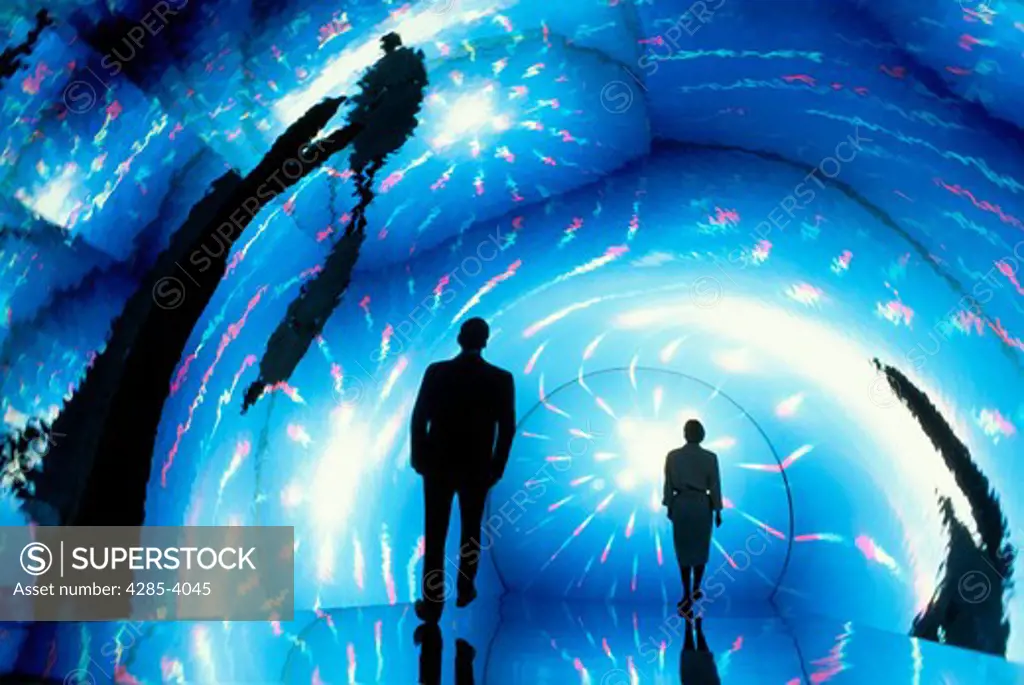Special effects shot of two people walking down a reflective tube illuminated by blue lights and prismatic bursts.