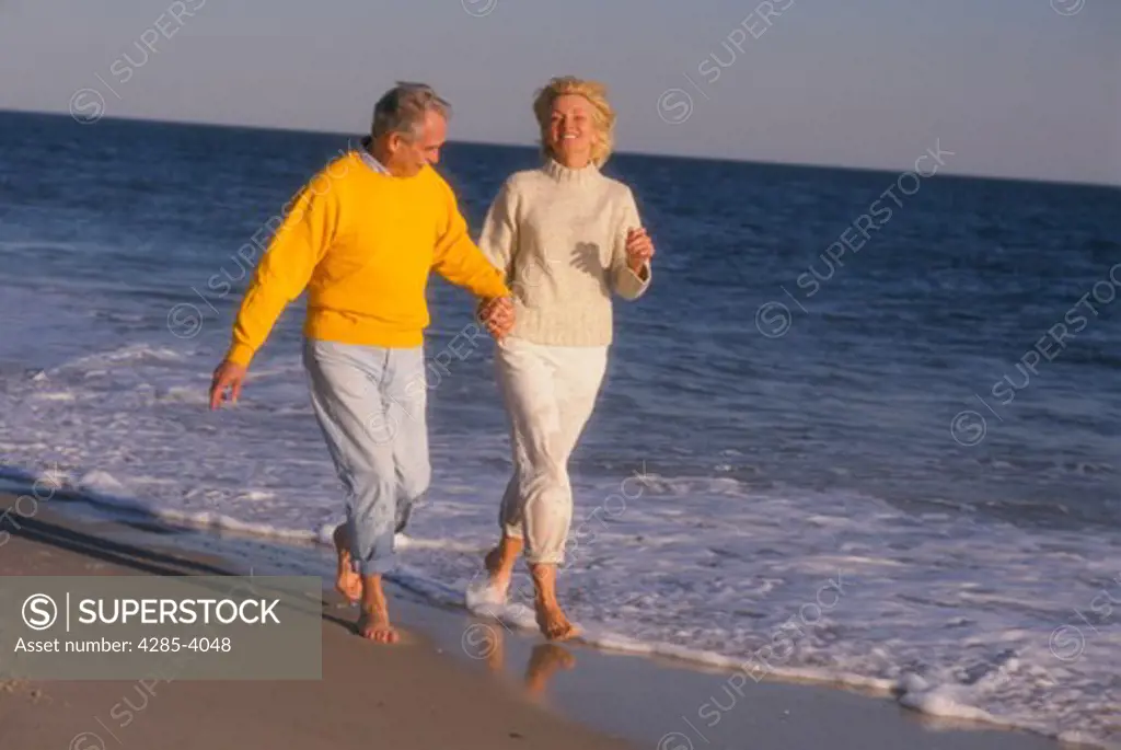 Barefoot middle-age couple wearing sweaters and rolled-up pants walking hand in hand along a beach.