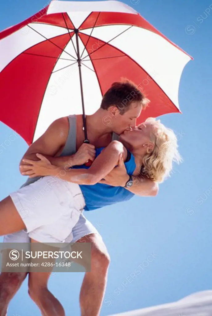 A man and woman kissing passionately in the shade of a red and white umbrella.
