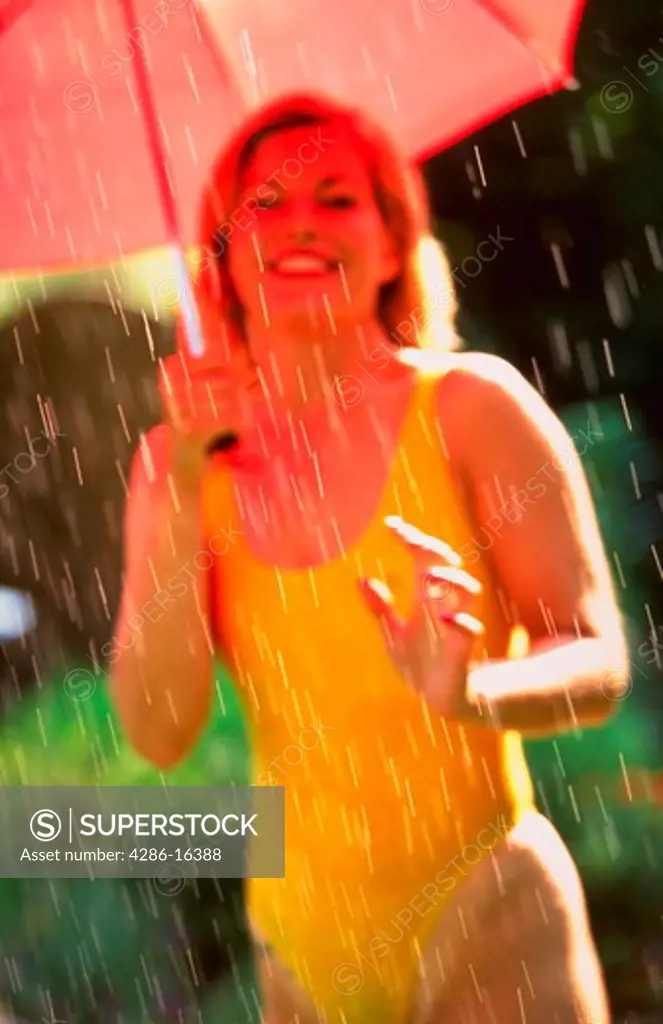 Woman in a yellow bathing suit holding a red umbrella while standing under the shower of a sprinkler.