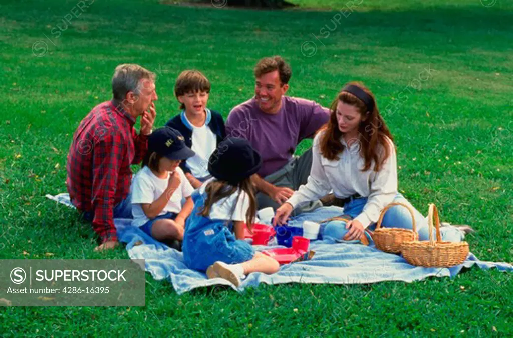 Three generations of a family with grandfather, parents and three children sitting on blanket having a picnic.
