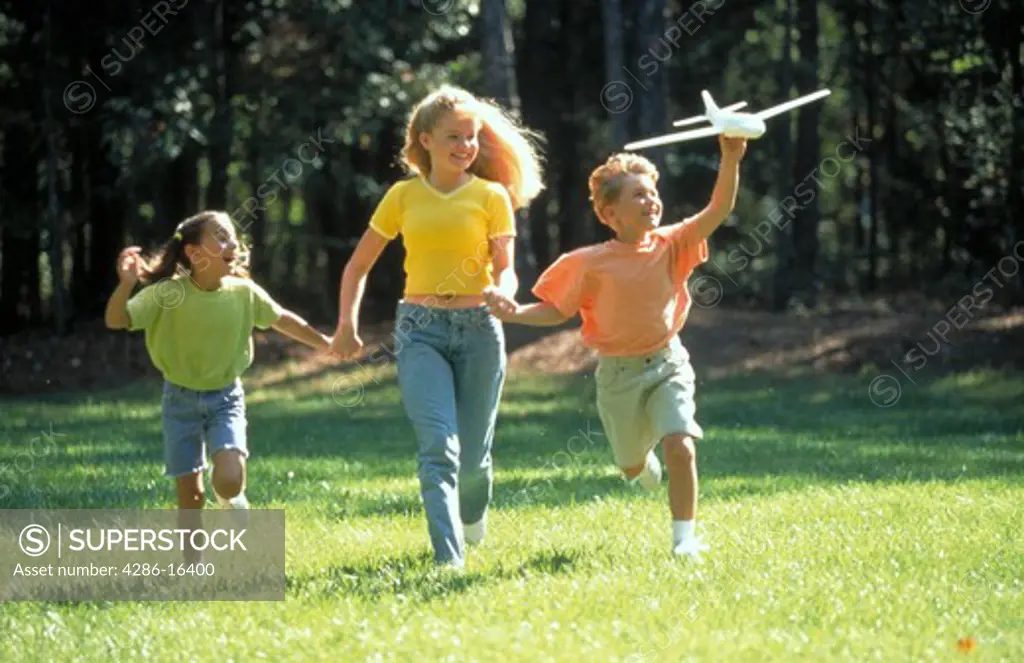Female teenager running through the grass with a young girl and a young boy who is holding a model airplane.