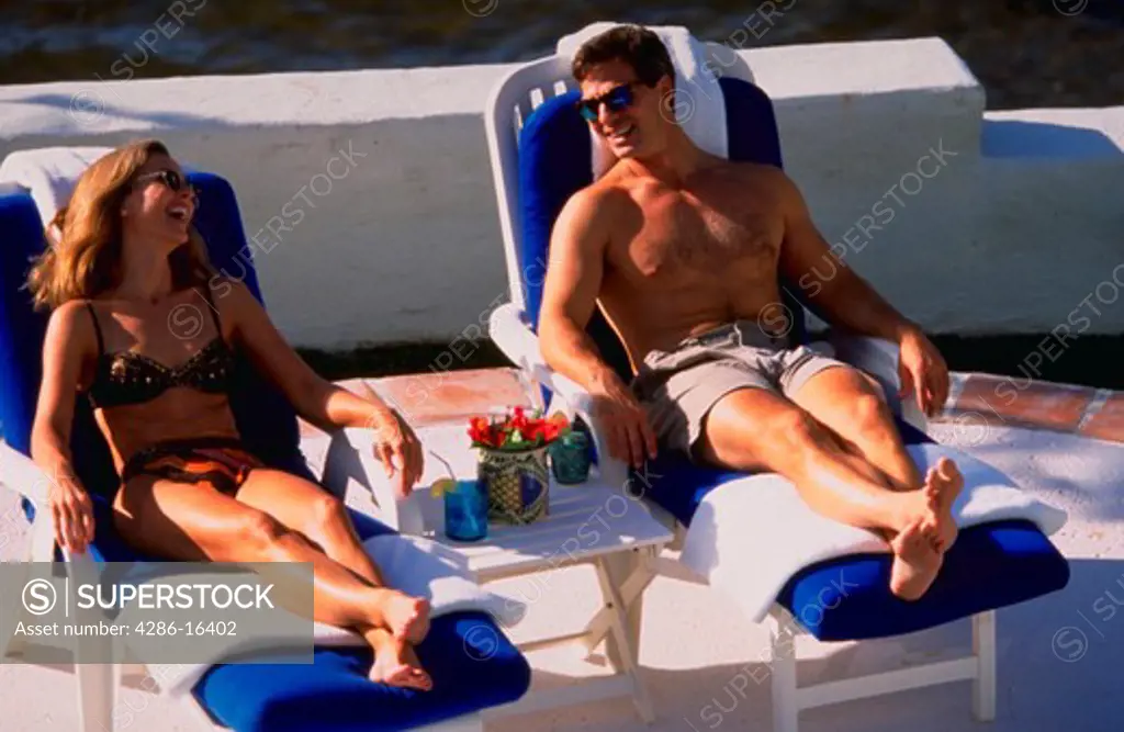 Man and woman wearing swimsuite and sitting on lounge chairs in the sun laughing.