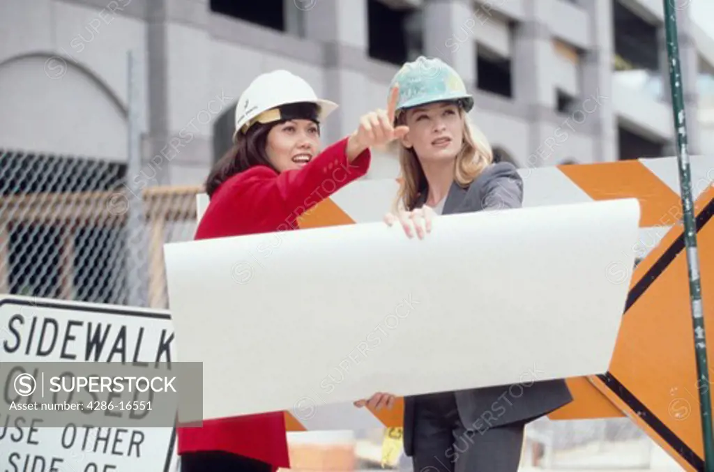 Two female architects holding blue prints and wearing hardhats while inspecting a work site.