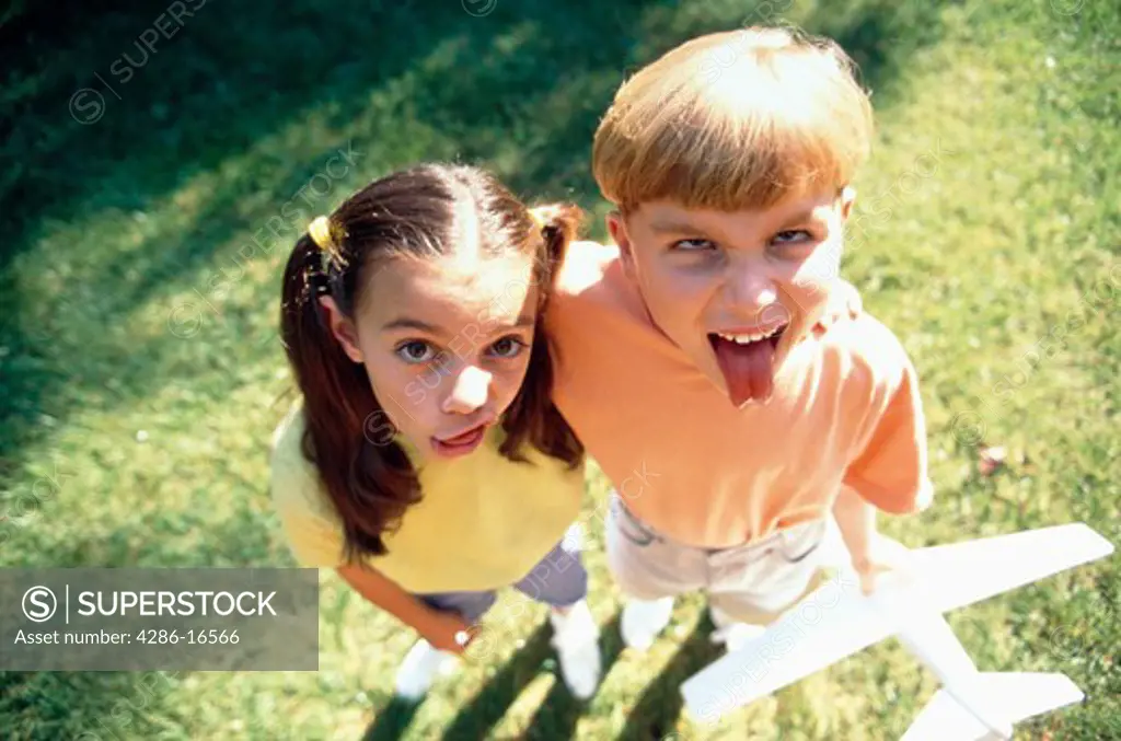 Aerial view of a young boy and girl making faces at the camera while holding a model airplane.