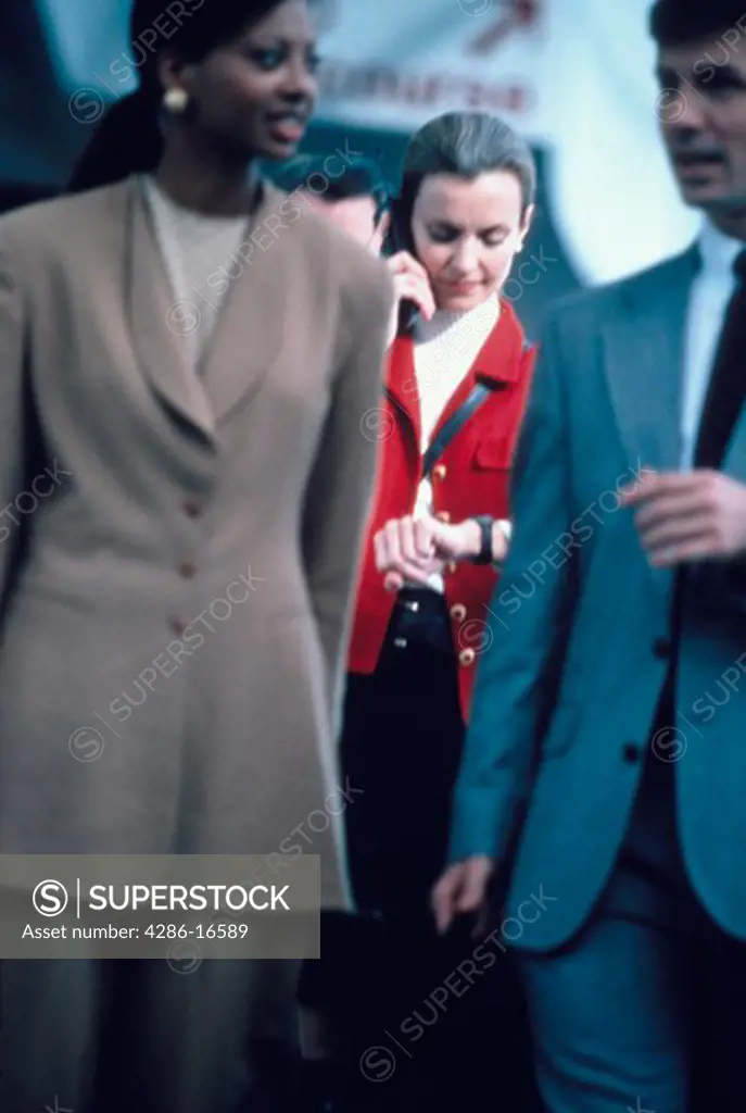 Two business people talking as a businesswoman standing behind them talks on a cellular phone and checks her watch.