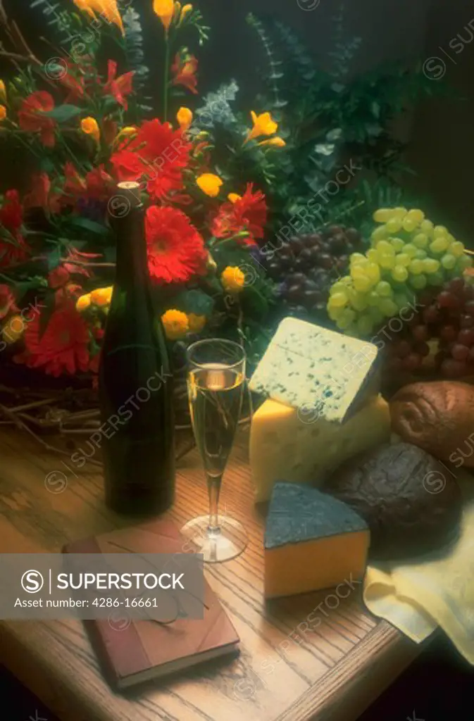 Still life of cheese, wine, grapes, bread, wine in a glass, a flower arrangement, and a pair of eyeglasses resting on a book.
