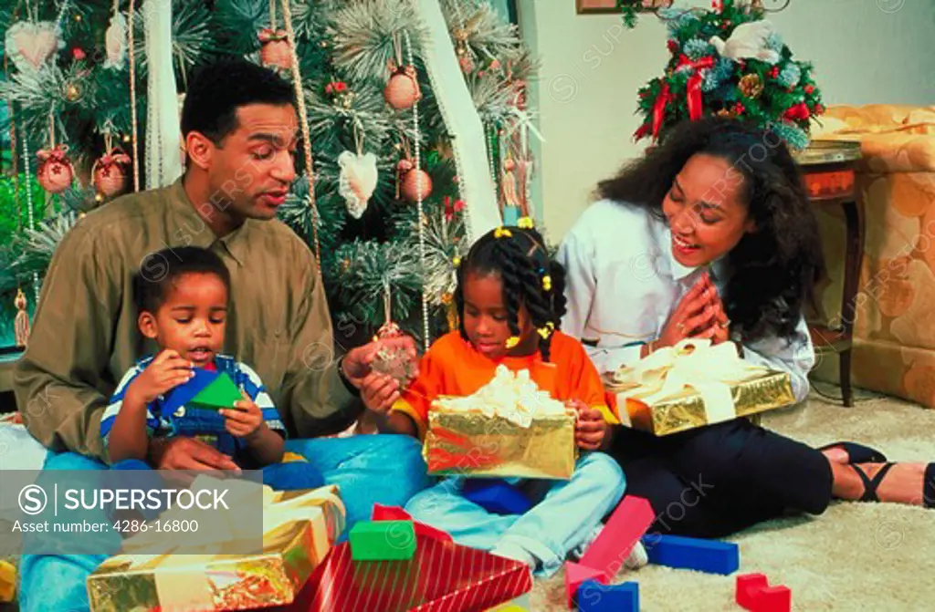 An African-American family, consisting of a mother, father, son and daughter, sitting on the floor next to a Christmas tree opening presents on Christmas morning.