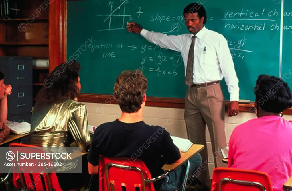 An African-American high school math teacher standing in front of a chalk board and pointing at a diagram on the board lecturing to his students.