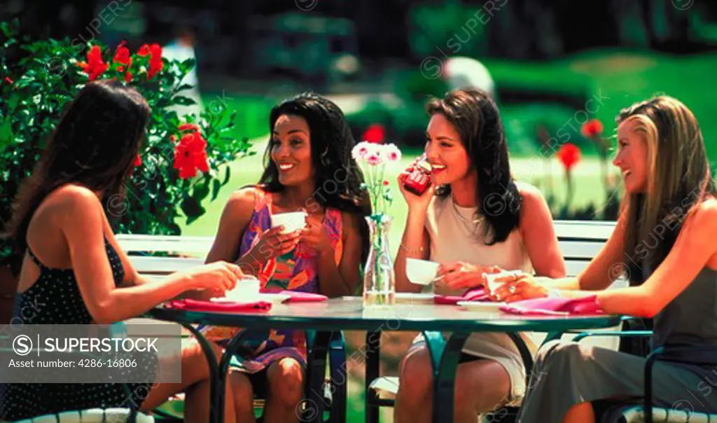 A group of four young women sitting outside at a table having coffee together and laughing. One woman is holding a cell phone.