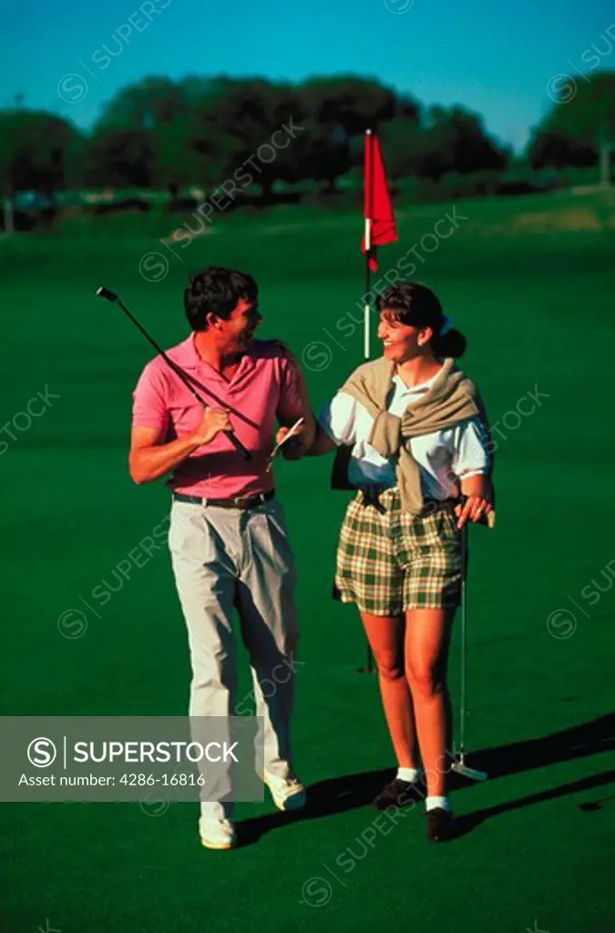 A smiling couple carrying golf putters walking across a putting green with the cup and flag in the background.