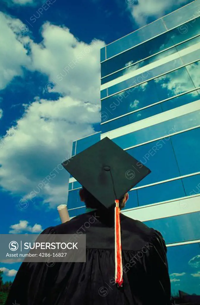 View from behind a college graduate wearing his cap and gown as he looks forward to a career in the business world. He is looking up at the exterior of a glass office building.