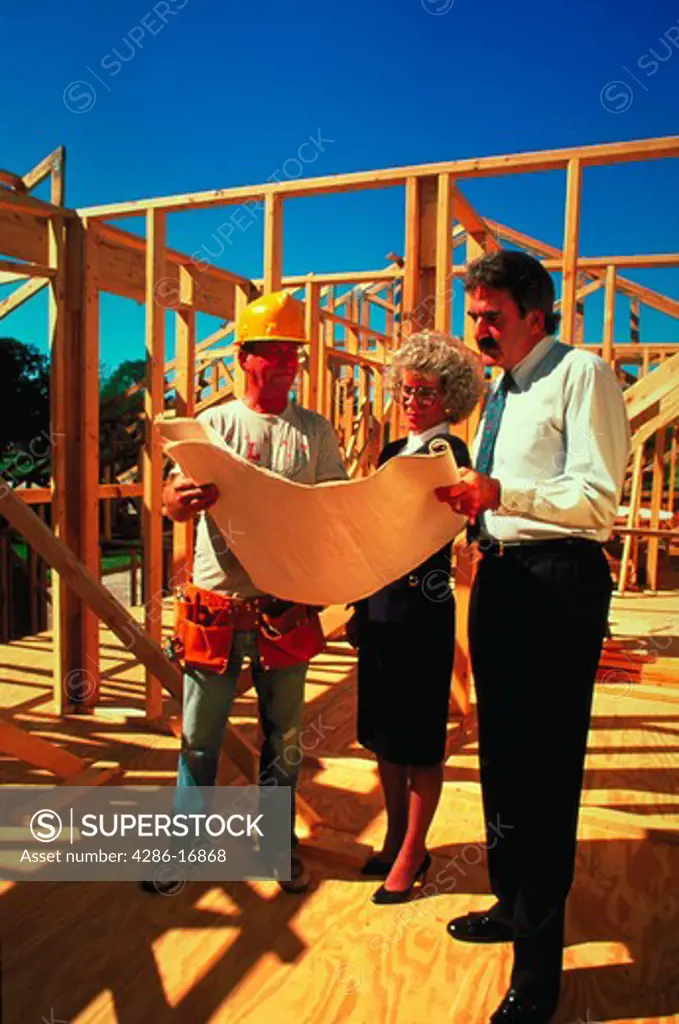 A man and a woman confer with a construction worker over blue prints while standing in a house that is under construction.