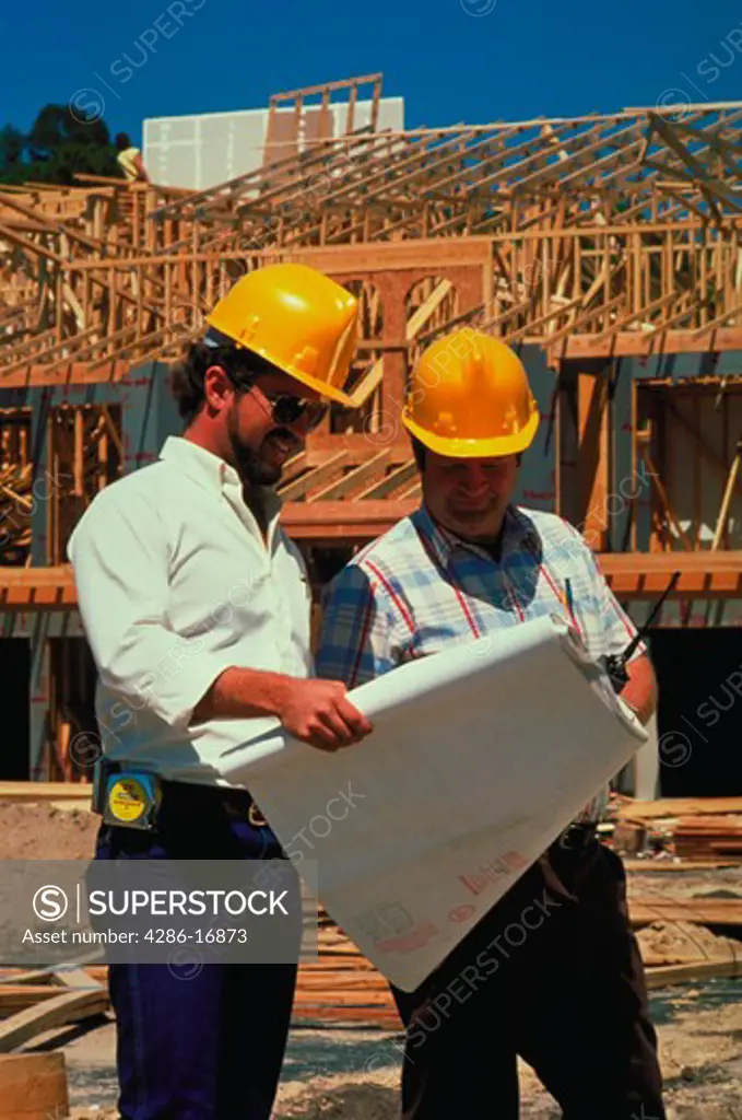 Two construction workers wearing yellow hard hats confer over blue prints with a building under construction in the background.
