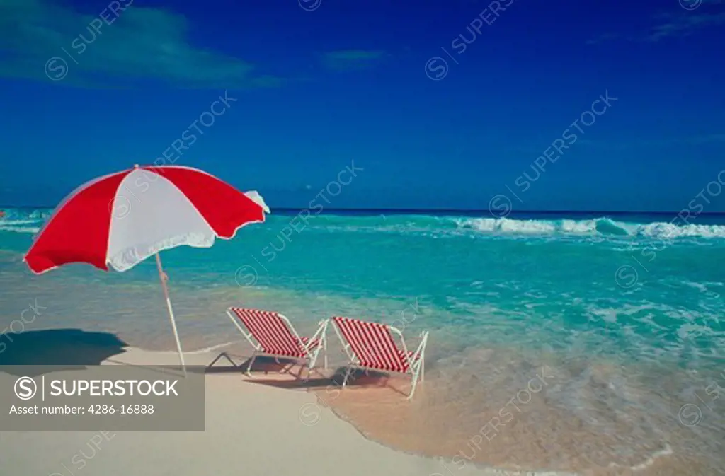 Two red an white chairs and a red and white umbrella sit just at the edge of the ocean in the sand beneath a blue sky.