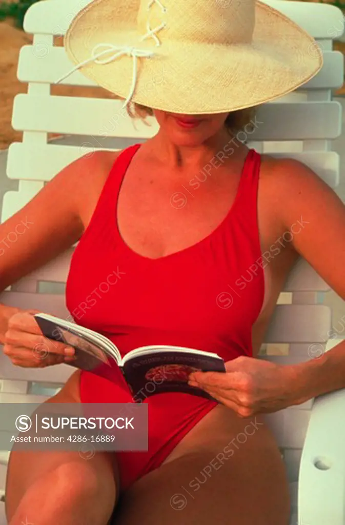 A woman wearing a red bathing suit and a wide brimmed hat relaxing on a lounge chair in the sun while reading a book.